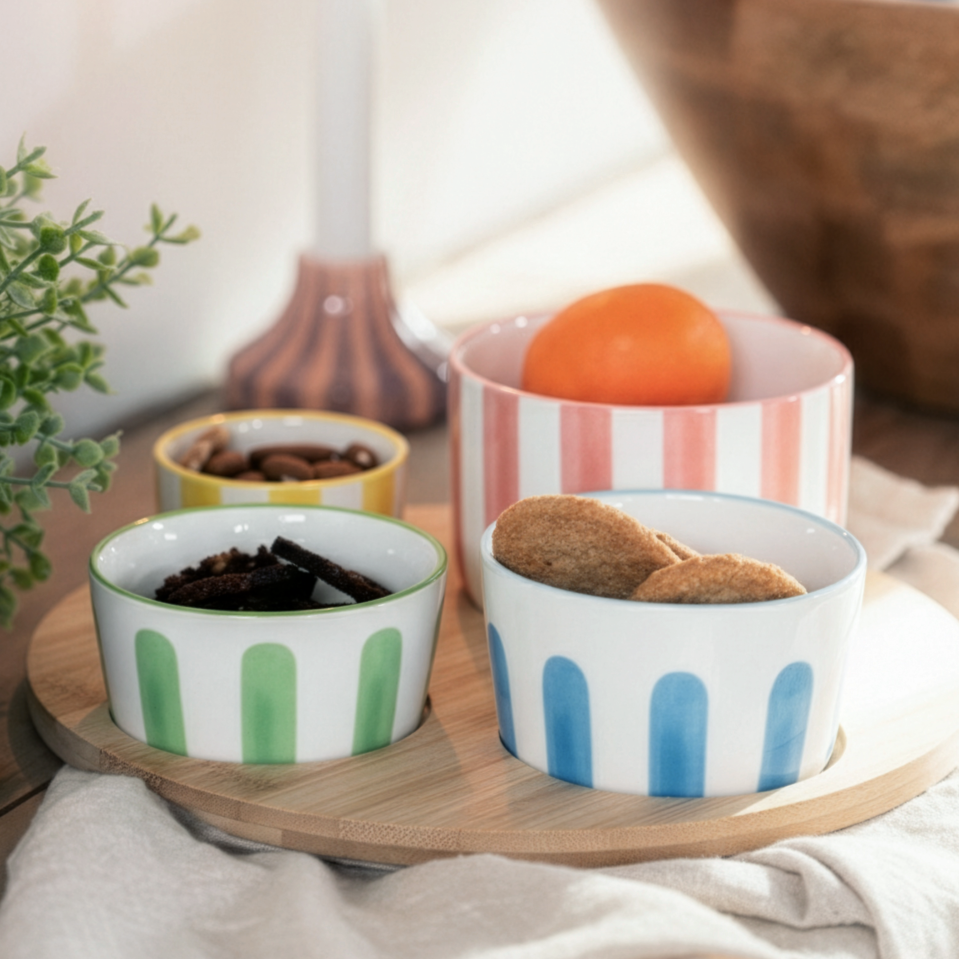 Small striped bowls with snacks on a wooden tray next to a candle and plant.